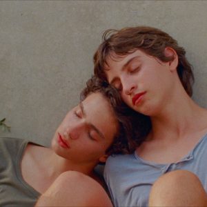 two boys sit with their backs to a wall, resting their heads on each other with their eyes closed